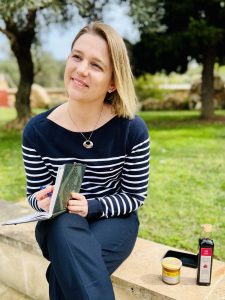 Jessica Deutsch sitzt nachdenklich mit einem Buch auf einer Mauer, neben ihr zwei hochwertige Marketingprodukte als Symbol für strategische Kundenbindung.